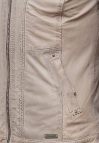 Beige läderjacka med en slät yta, silverfärgad dragkedja och en ficka med tryckknapp. Har sömnadsdetaljer och en märkesetikett.