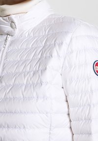 Vit puffjacka med hög krage, vadderad struktur, dragkedja fram och en cirkulär logotyp på överärmen. Mjuk tygfinish.