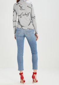 Šedá dlouhá mikina s černým kurzívním textem, v kombinaci se světle modrými skinny džíny a červenými sandály na vysokém podpatku. Jasné detaily látky a designu jsou viditelné.