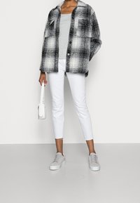 Sopra una canotta grigia, una shacket a quadri bianco e nero, abbinata a pantaloni bianchi cropped e sneakers grigio chiaro. Gli accessori includono una borsa bianca.