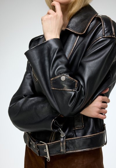 Black faux leather jacket with a wide collar, silver hardware, and contrasting stitching. Features zip pockets and a belted waist.