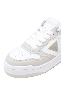 Weiße und hellgraue Guess-Sneaker mit perforierter Zehenspitze, weißen Schnürsenkeln und dreieckigem Guess-Logo an der Seite.