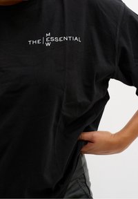 Svart bomulls-t-shirt med vit text "THE ESSENTIAL M W". Kort ärm, avslappnad passform och slät textur. Hand placerad på sidan.