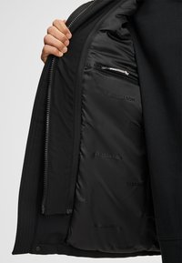 Czarna kurtka o gładkiej powierzchni zewnętrznej. Podszewka wewnętrzna ozdobiona subtelnym wzorem z logo. Posiada kieszeń na zamek oraz solidne zapięcie na zamek błyskawiczny.