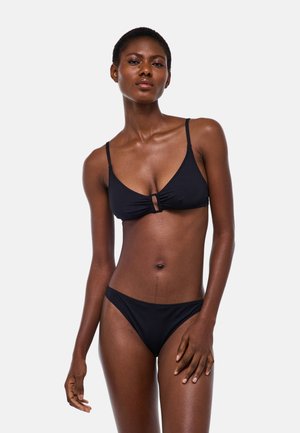 Femme portant un bikini noir avec une petite découpe devant sur le haut, debout devant un fond gris clair uni.