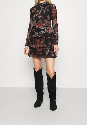 Mujer con vestido mini negro de manga larga con estampado floral, falda con capas y botas negras de ante hasta la rodilla sobre un fondo liso.