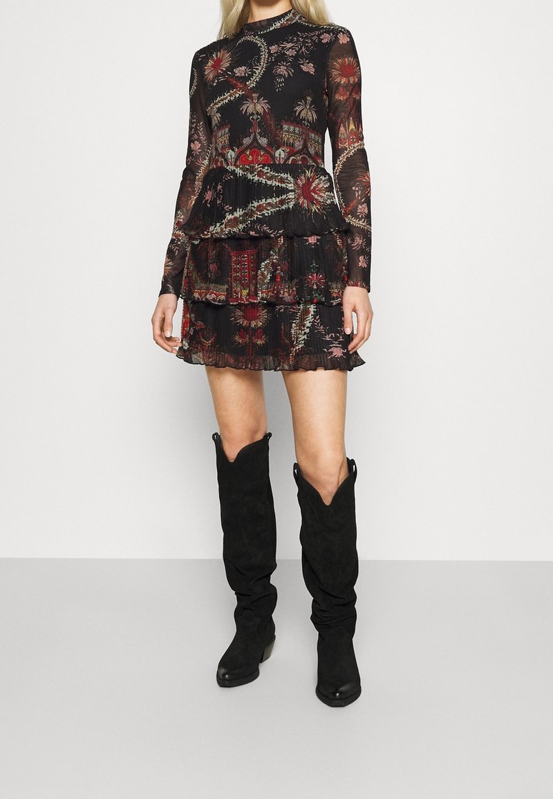 Femme portant une mini-robe noire à manches longues avec imprimé floral, jupe à volants et bottes hautes en daim noir sur fond uni.