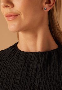 Runde Steckerohrringe mit einem hellblauen Edelstein, in Silber gefasst. Das Model trägt ein strukturiertes schwarzes Oberteil, das die Ohrringe hervorhebt.