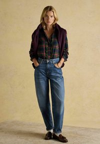 Plaid button-up shirt in green, red, and yellow; dark purple sweater draped over shoulders; high-waisted blue jeans and brown moccasins.