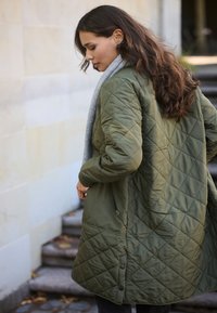 Giacca trapuntata verde con vestibilità rilassata, dotata di una sciarpa grigia, tasca laterale con zip e dettagli a pressione all'orlo.