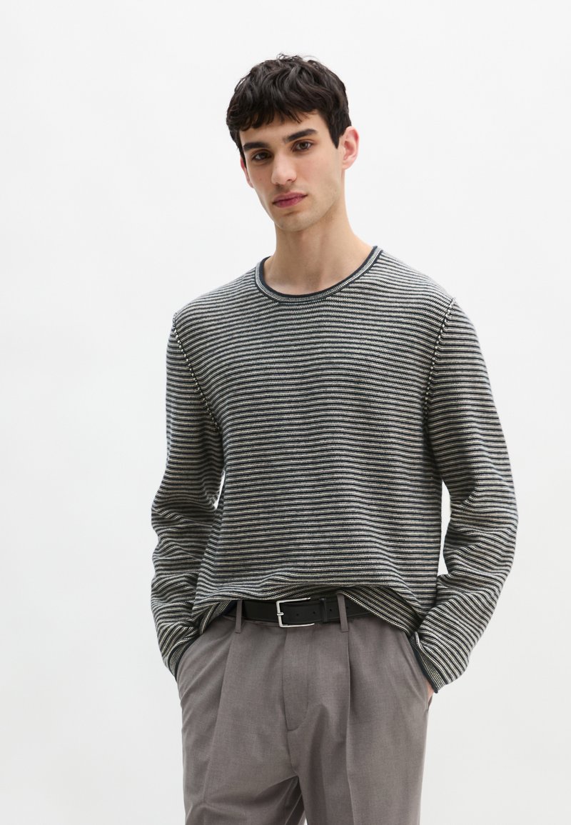 Gestreifter Pullover in Schwarz und Weiß, mit Rundhalsausschnitt und lockerem Schnitt. Das Modell kombiniert ihn mit grauen Hosen und einem schwarzen Gürtel und zeigt so einen lässigen Look.