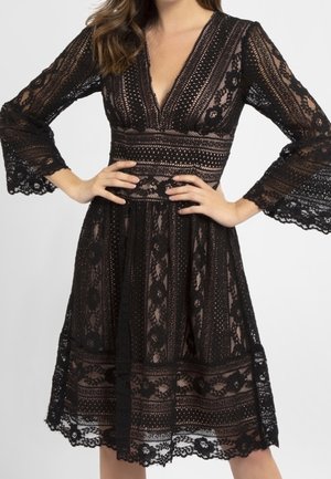 Femme portant une robe en dentelle noire avec un profond décolleté en V, des manches trois-quarts évasées et une taille cintrée, debout les mains sur les hanches.