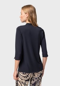 Top in poliestere blu navy con scollo tondo e maniche a 3/4. Texture liscia e design semplice, abbinato a pantaloni beige con fantasia.