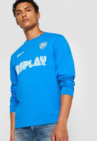 Ung mand iført en lys blå langærmet skjorte med "REPLAY" logo og skjold, iført blå jeans, poserende mod en ensfarvet lys baggrund.