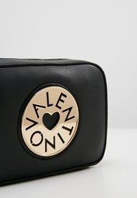 Musta suorakulmainen laukku, joka on valmistettu teksturoidusta nahasta. Sen etupuolella on pyöreä kultaembleemi, jossa on leikatut kirjaimet ja sydämen muoto.