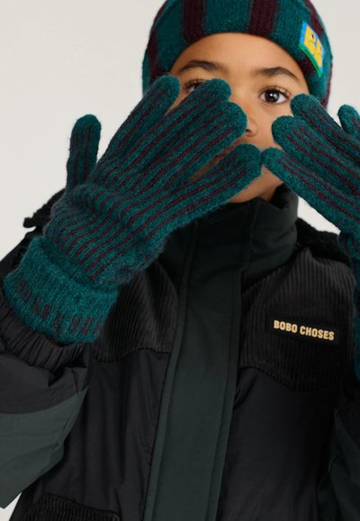 Enfant portant une veste noire et un bonnet et des gants tricotés rayés verts et bordeaux assortis, tenant les mains près du visage.