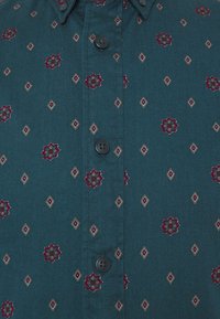 Teal skjorta med knappar som har ett mönster av röda blommor och diamanter, med subtila gröna accenter och svarta knappar.