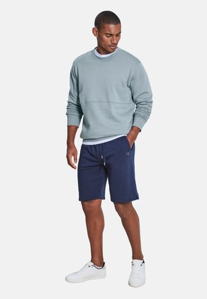 Mann trägt hellblauen Pullover, marineblaue Shorts mit Kordelzug und weiße Turnschuhe, steht mit einer Hand in der Tasche und schaut nach unten.