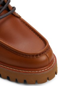 Bota de cuero marrón con puntera redondeada, costuras resistentes y una suela de goma texturizada con tacos pronunciados. Cordones grises pasados por ojales.