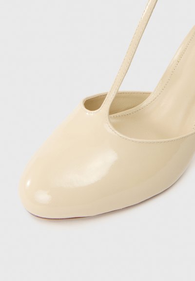 Botte à talon haut en cuir verni beige avec un bout arrondi et une seule sangle de cheville. Fini lisse et brillant avec des détails de couture visibles.