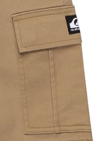 Poche cargo beige en tissu durable avec des coutures visibles. Elle possède une fermeture à rabat et une étiquette de logo noire sur le côté.