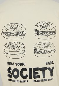 Krémově zbarvená látka s ilustracemi čtyř bagelů obrysovaných černě, s různými posypy. Text: "NEW YORK BAGEL SOCIETY RUCNĚ VÁLENÉ BAGELY."