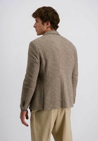 Brauner strukturierter Blazer mit Reverskragen, einem Schlitz und knöpfbaren Manschetten, kombiniert mit hellen Hosen.