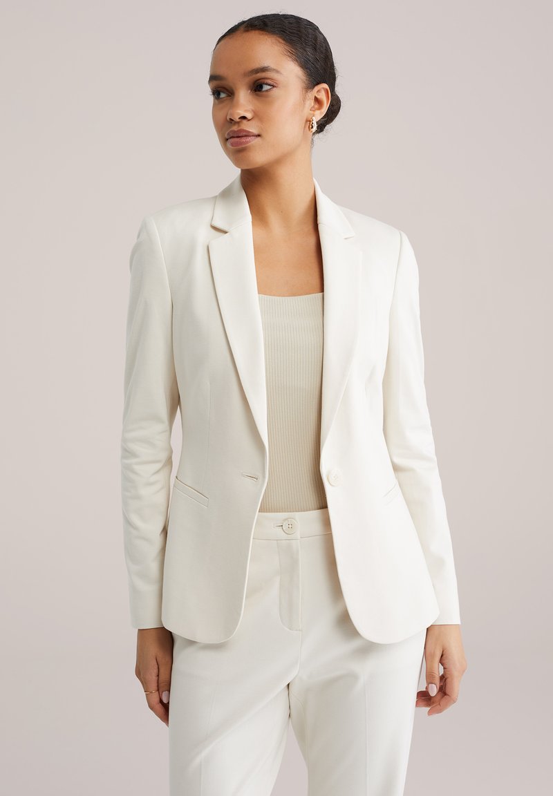 Moteris su tamsiais plaukais, surištais žemai kuodu, dėvi auksinius apskritus auskarus, kreminės baltumo švarką, derančias kelnes ir ruduotas smėlio spalvos megztinį, stovinčią neutraliame fone.