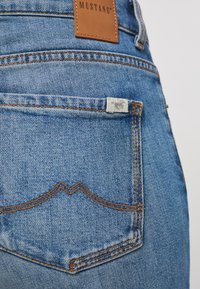 Jeans de mezclilla azul claro con un bolsillo trasero que presenta un diseño de costura curvada y una pequeña etiqueta de marca. Tejido texturizado con costuras visibles.
