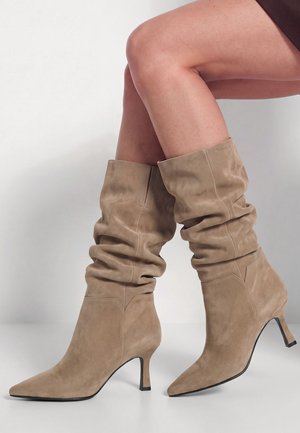 Modèle portant des bottes hautes en daim beige à l’aspect souple et plissé, avec des bouts pointus et des talons fins, sur un fond uni.