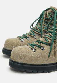 Solidne beżowe buty trekkingowe o chropowatej powierzchni, z zielono-beżowymi sznurowadłami na czarnych, grubych podeszwach.