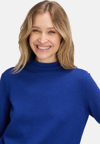 Blå polotröja i mjukt tyg, med långa ärmar och en figursydd design, har en slät textur och inga synliga mönster.