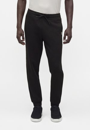 Bărbat purtând pantaloni de trening negri cu șnur și adidași negri cu talpă albă, stând în picioare în fața unui fundal simplu, deschis.