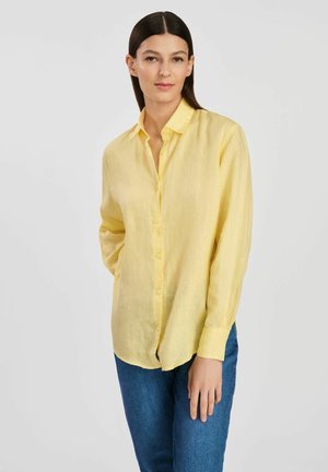 Żółta lniana koszula zapinana na guziki z długimi rękawami, z klasycznym kołnierzykiem i luźnym krojem, w połączeniu z niebieskimi dżinsami.