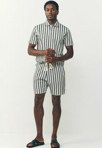 Chemise à rayures à manches courtes et shorts assortis en marine et crème, avec un col, une fermeture à boutons et une taille à cordon. Sandales noires à enfiler.