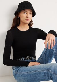 Zwart shirt met lange mouwen van lichtgewicht materiaal, gecombineerd met blauwe spijkerbroek en een zwarte geribbelde bucket hat. Zwarte riem met zilveren gespen.