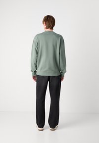 Una persona è in piedi di spalle, indossa una felpa verde a maniche lunghe, jeans neri e scarpe bianche, su uno sfondo bianco uniforme.