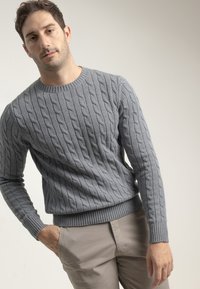 Suéter gris de punto trenzado con cuello redondo y puños y dobladillo acanalados, combinado con pantalones beige claro.
