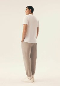 Weißes schlichtes T-Shirt mit kurzen Ärmeln, graue taillierte Jogginghose und weiße Sneaker. Das Model steht mit dem Rücken zur Kamera, die Hände in den Taschen, vor einem schlichten Hintergrund.