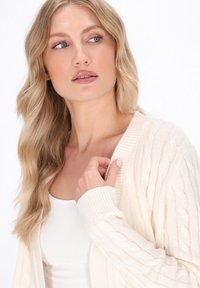 Cable-knit, creme cardigan med ribbede manchetter og krave; letvægts, tekstureret stof. Modellen har en tætsiddende hvid top på under.