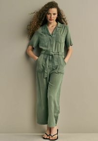 Zelený džínový overal s krátkymi rukávmi, dve predné vrecká, opasok v páse a široké nohavice. Čierne sandále na opätku dopĺňajú outfit.
