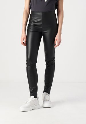 Person trägt schwarze, hoch taillierte, eng anliegende Lederhose und weiße klobige Sneakers, steht vor einem schlichten hellen Hintergrund.