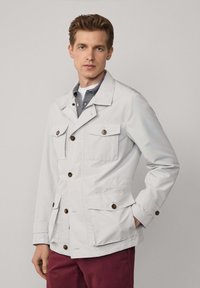 Hellgraue äußere Jacke mit strukturiertem Design, vier Fronttaschen, braunen Knöpfen und glatter Textur. Über einem geschichteten Hemd getragen.