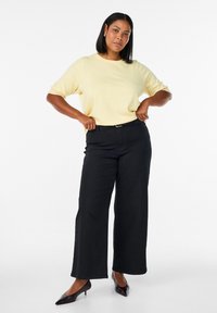 Gelber Pullover mit kurzen Ärmeln, kombiniert mit schwarzen, weit geschnittenen Hosen und spitzen schwarzen Absätzen. Einfaches Design mit entspanntem Sitz.