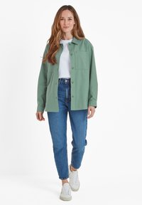 Green button-up jacket, two chest pockets, worn over a white shirt, paired with blue jeans and white sneakers. Casual, relaxed fit.