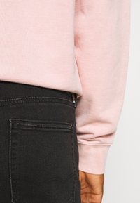 Sweat-shirt rose clair associé à un jean noir. Le sweat-shirt présente un design simple avec une texture lisse, tandis que le jean comporte des détails de couture et une poche arrière.