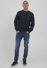 Tummansininen crewneck-huppari pehmeästä kankaasta, yhdistettynä sinisiin farkkuihin ja mustiin tennareihin, luoden rentoa tyyliä.
