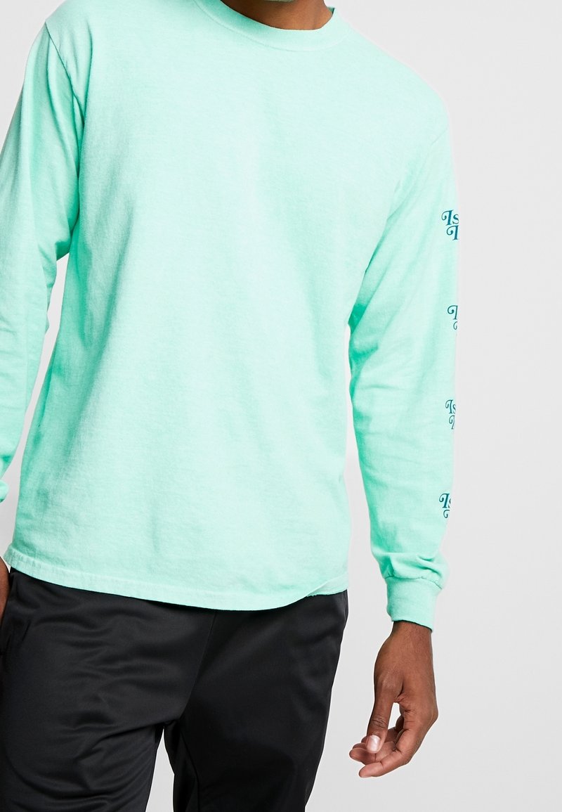 Minta vihreä pitkähihainen paita, joka on tehty pehmeästä kankaasta. Siinä on pyöreä pääntie ja tulostetut tekstit korostavat molempia hihoja. Yhdistetty mustiin housuihin.