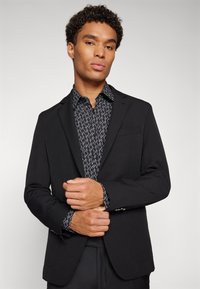 Sacou negru cu un design slim fit, închidere cu un singur buton și rever clasic, purtat peste o cămașă șteptată neagră și albă.