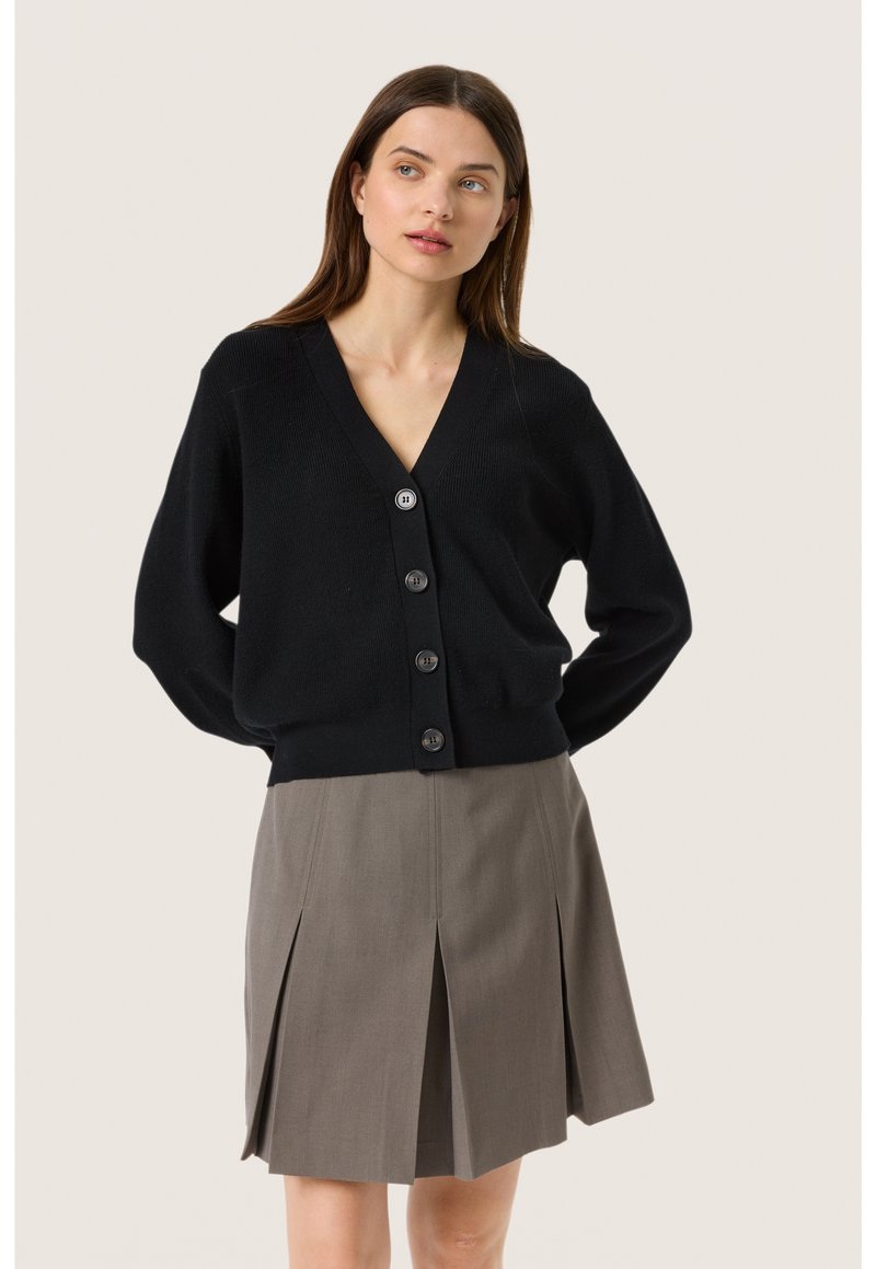 Svart stickad cardigan med V-hals, med knappstängning och långa ärmar, bärs över en grå plisserad kjol.